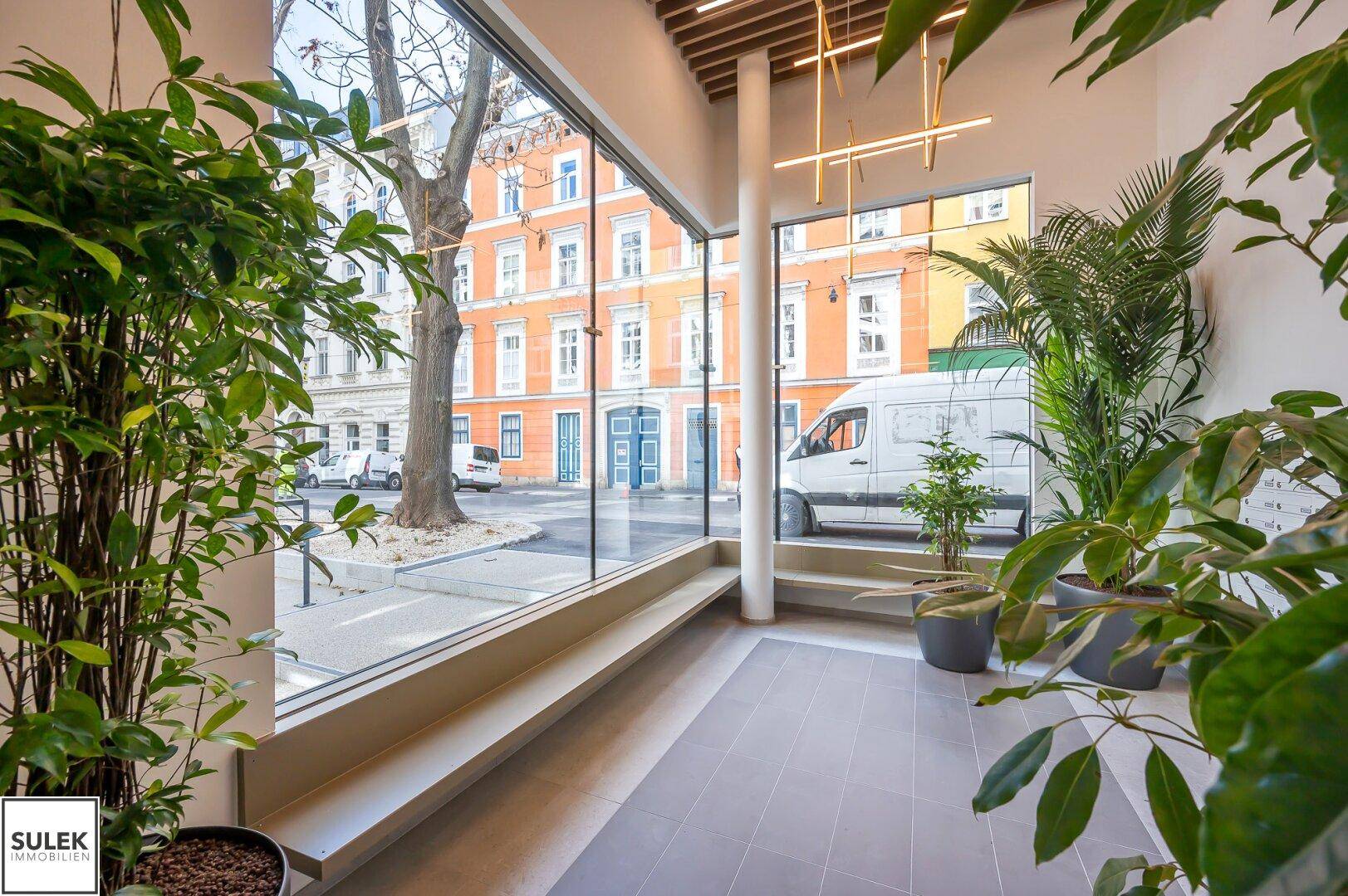 Moderner Eingangsbereich mit großen Glasfronten, Pflanzen und Blick auf die Straße.