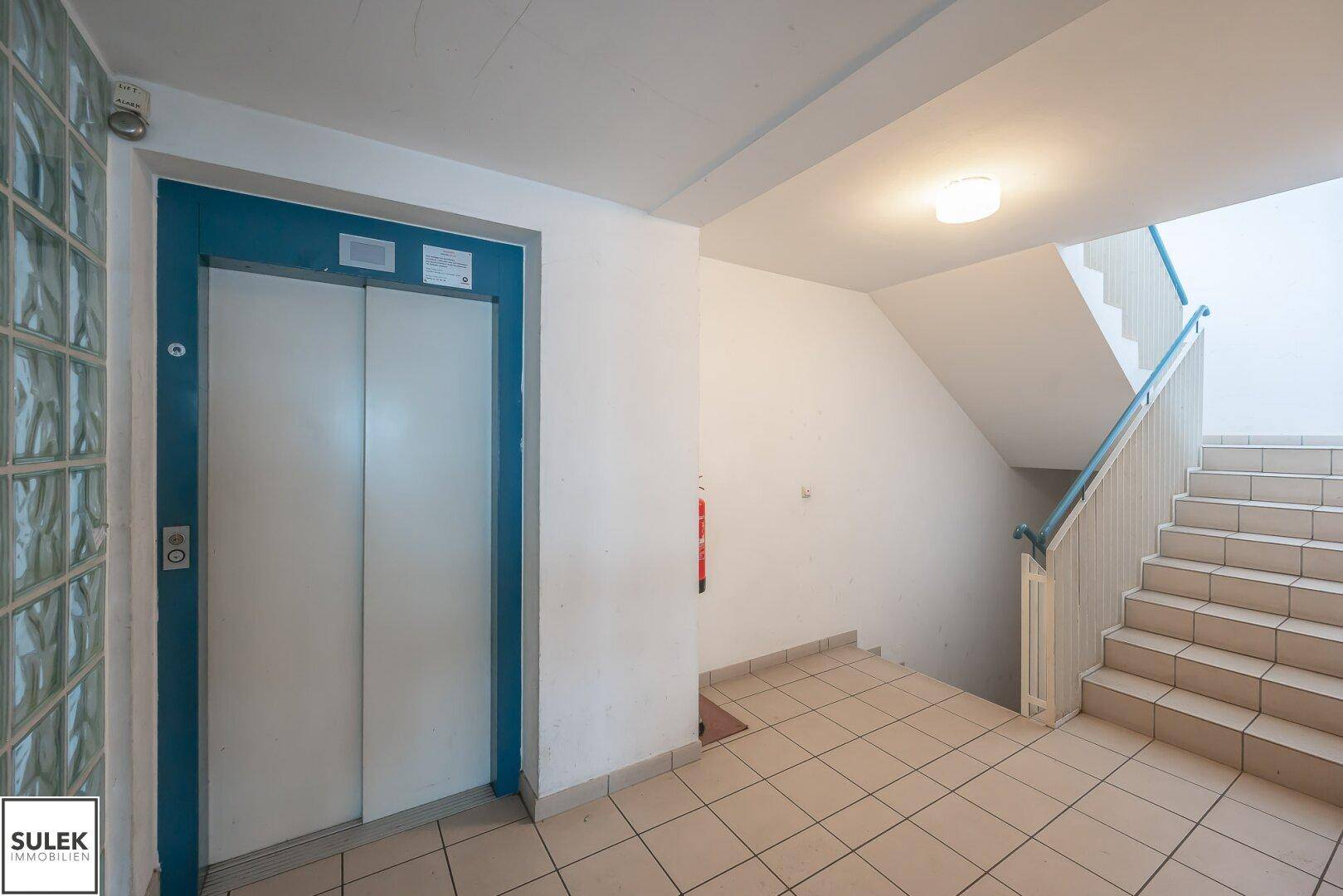 Eingangsbereich mit Aufzug und Treppenhaus, gefliestem Boden und weiß gestrichenen Wänden.