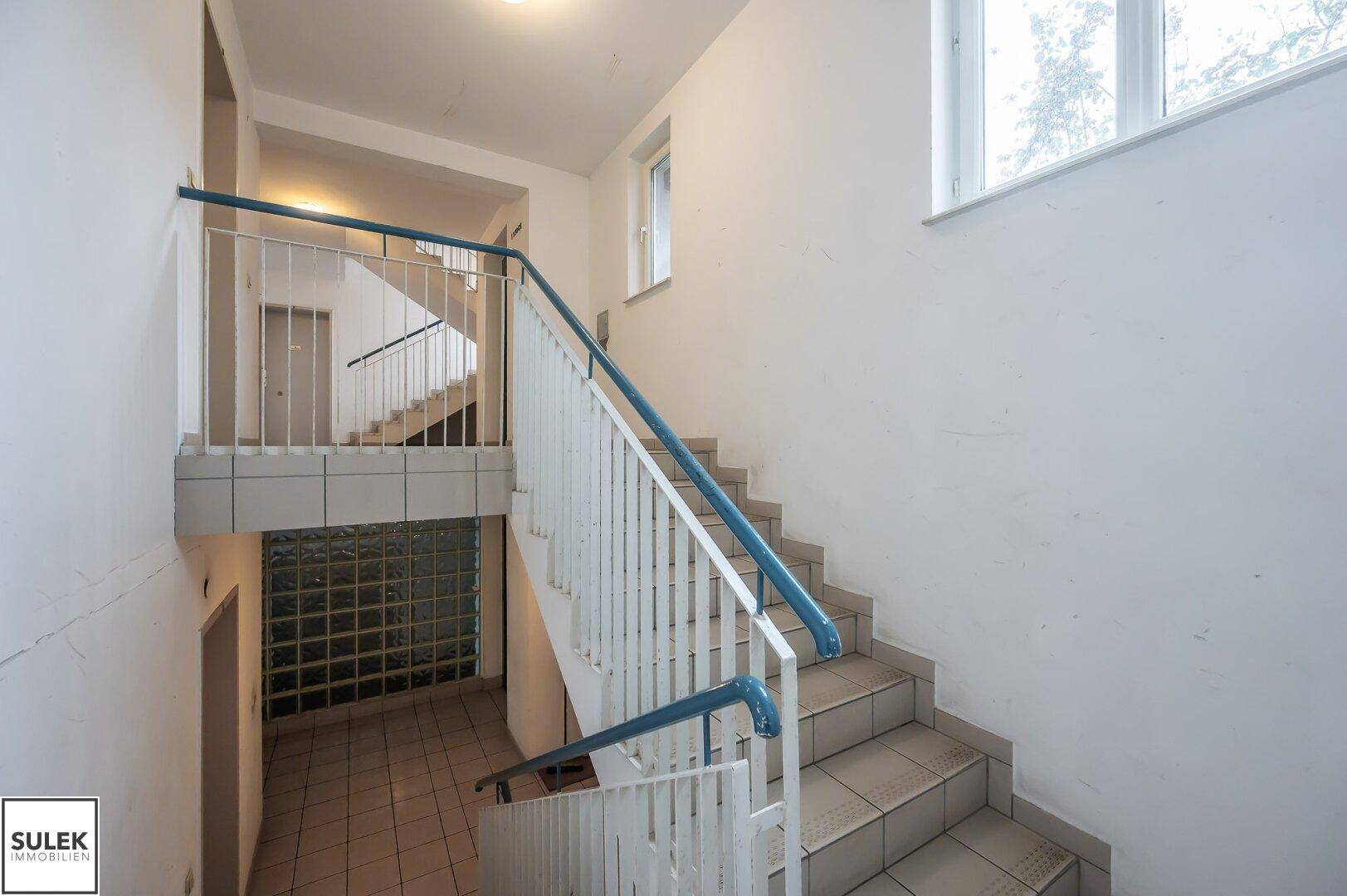Treppenhaus mit Fliesenboden, blauen Geländern und einem Fenster für natürliches Licht.