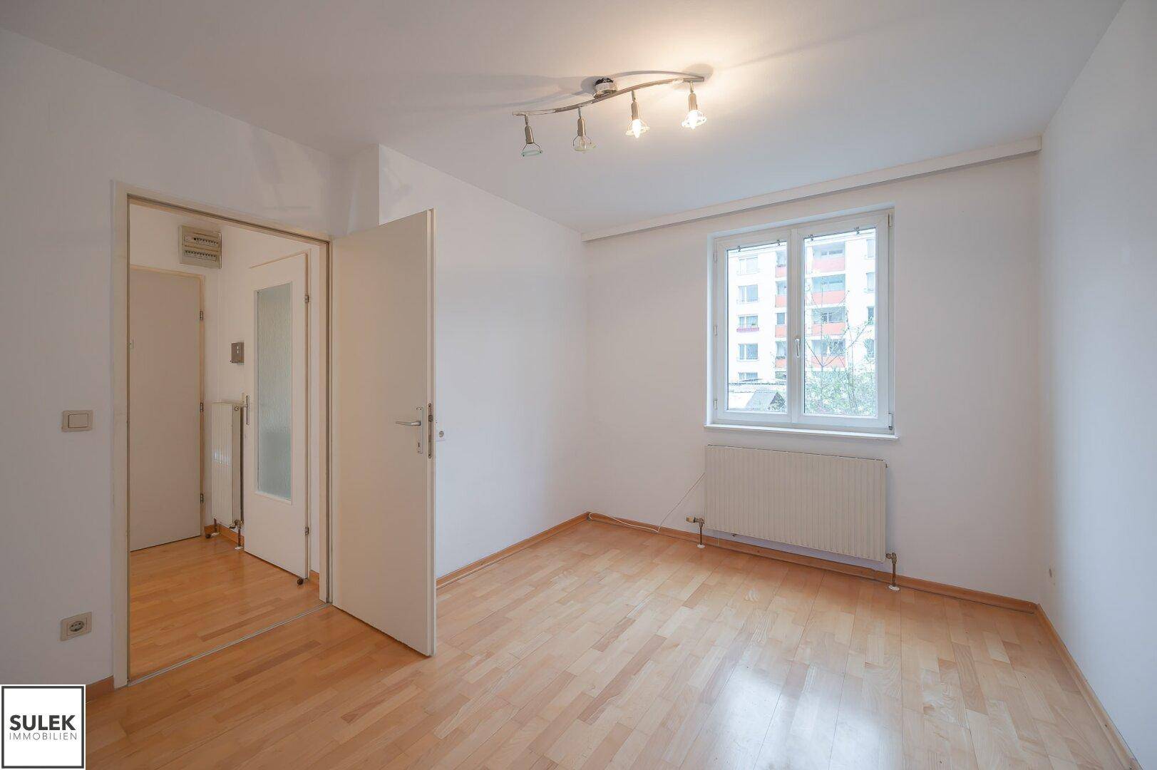 Leeres Zimmer mit hellem Parkettboden, weiß gestrichenen Wänden und einem Fenster.