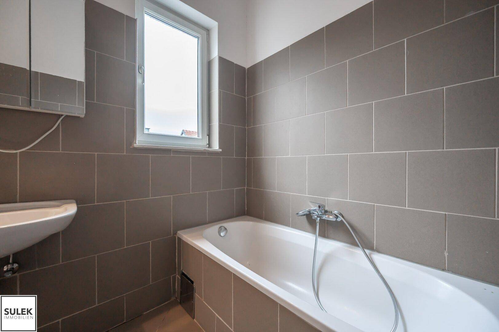 Badezimmer mit Badewanne, Fenster und grauen Fliesen an Wänden und Boden.
