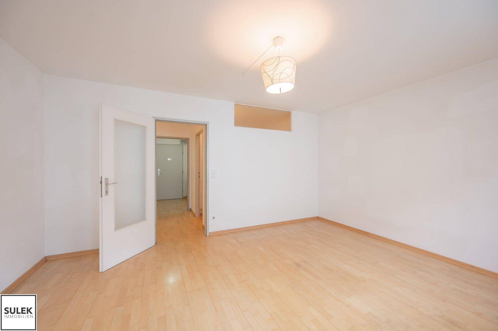 Helles, leeres Zimmer mit Parkettboden und einer Tür mit Glaseinsatz.