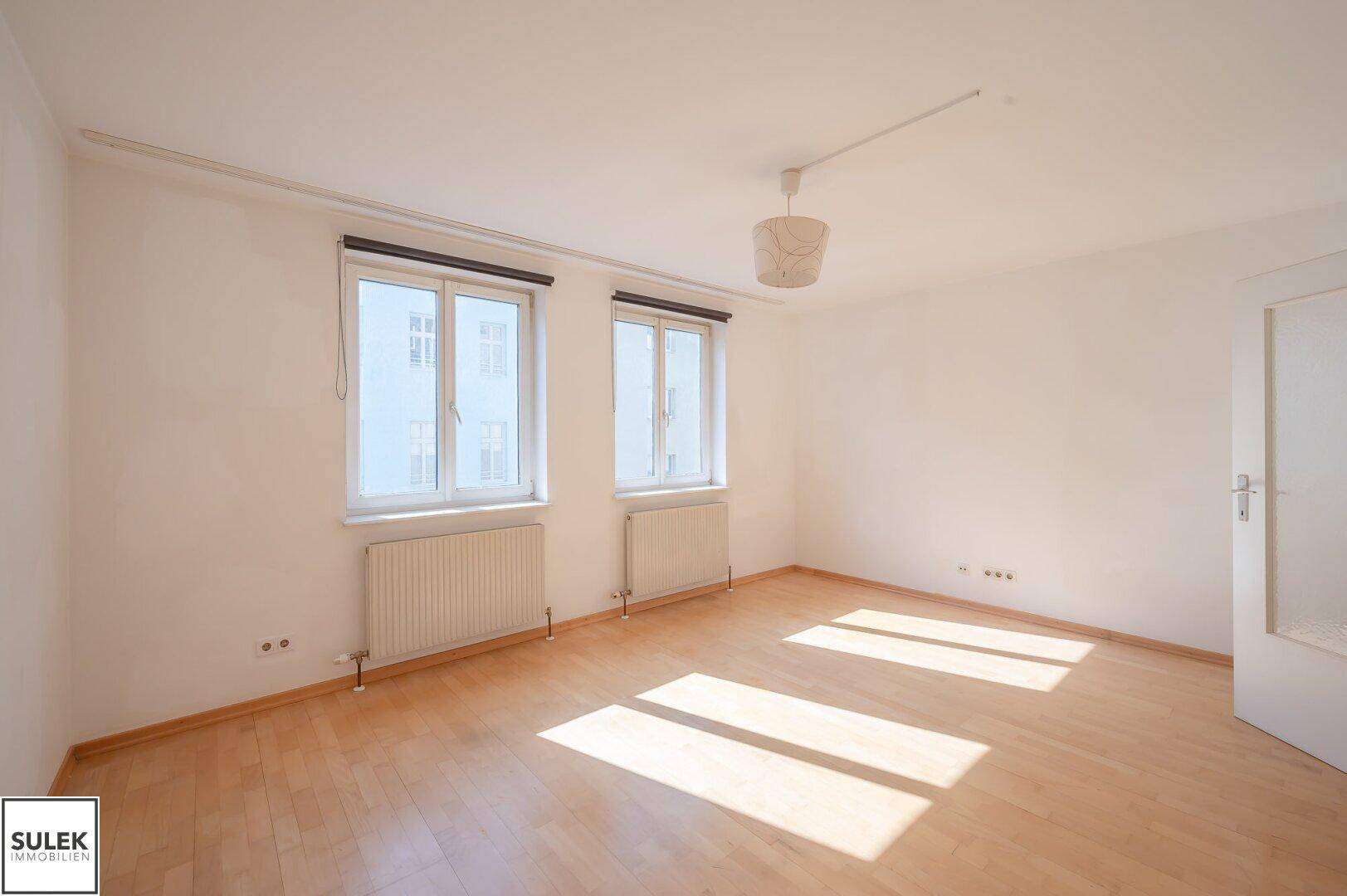 Lichtdurchflutetes Zimmer mit Parkettboden, zwei Fenstern und Heizkörpern darunter.