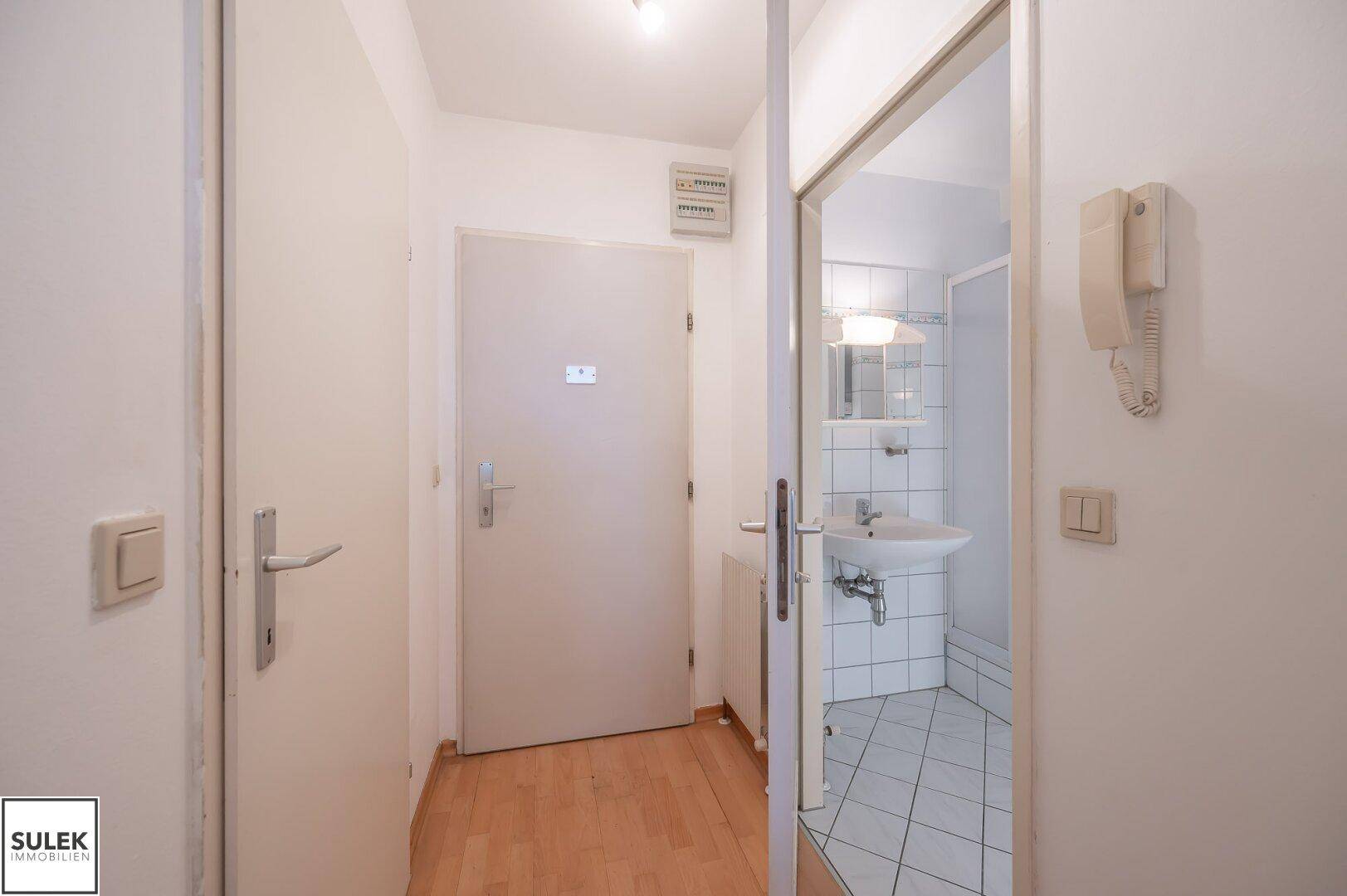Flur mit Holzboden und Blick in ein kleines Badezimmer mit Dusche und Waschbecken.