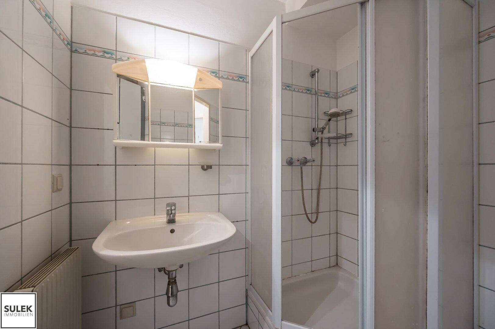Kleines Badezimmer mit Duschkabine, Waschbecken und weißen Fliesen an den Wänden.