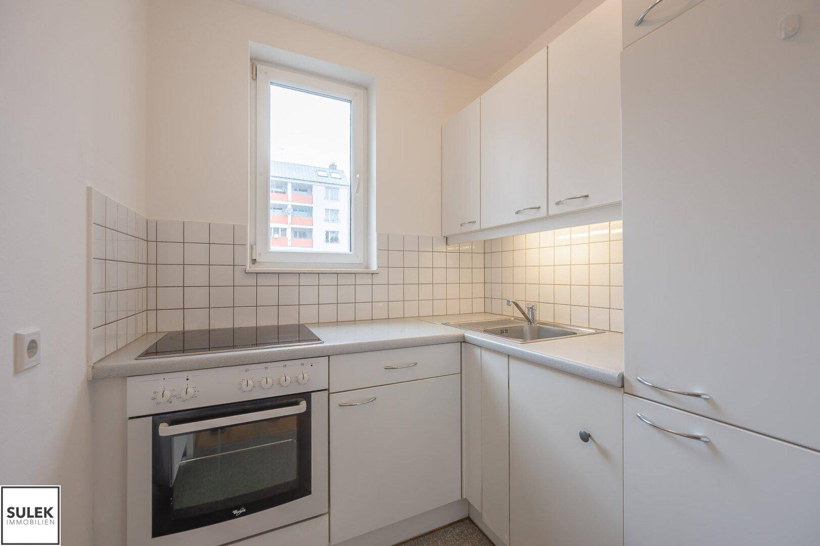 Helle Küche mit weißen Einbauschränken, Kochfeld, Backofen und einem Fenster.