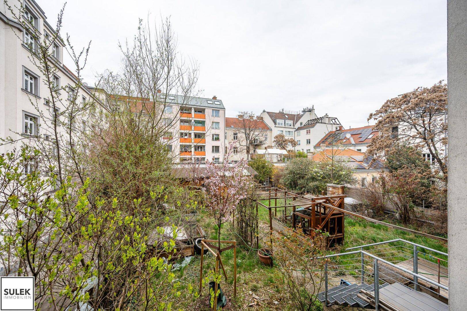 Blick auf einen grünen Hinterhofgarten mit Bäumen, Sträuchern und einer kleinen Treppe.