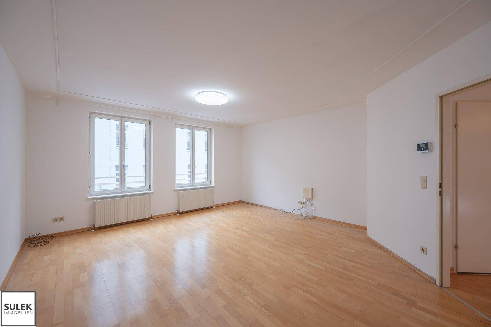 Großes Wohnzimmer mit zwei Fenstern und hellem Parkettboden.
