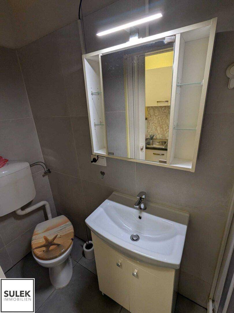 Kleines Badezimmer mit WC, Waschbecken und Spiegelschrank, komplett in Grau gefliest.