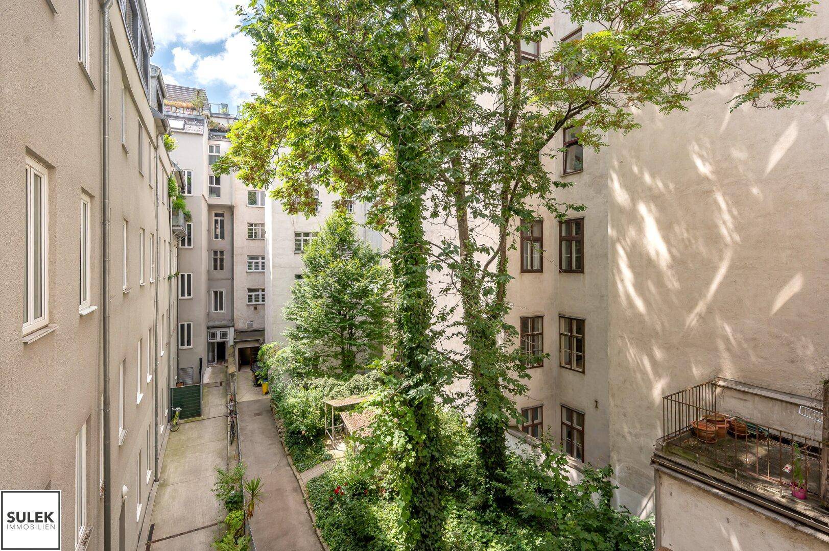 Blick auf den begrünten Innenhof mit hohen Bäumen und umliegenden Gebäuden.