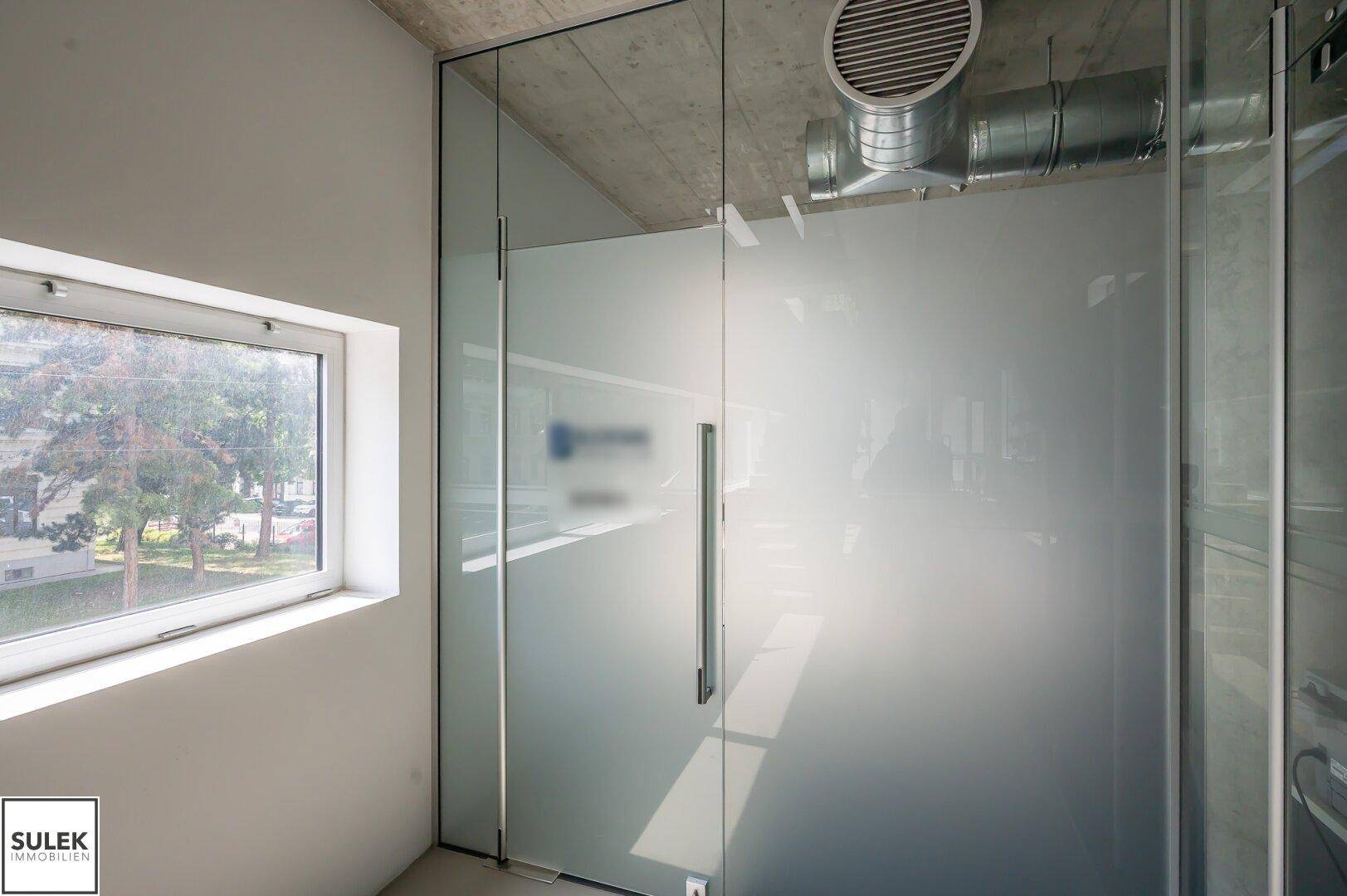 Innenansicht mit Milchglas-Trennwänden, einem Fenster und einer Sichtbetondecke mit Lüftungsrohren.