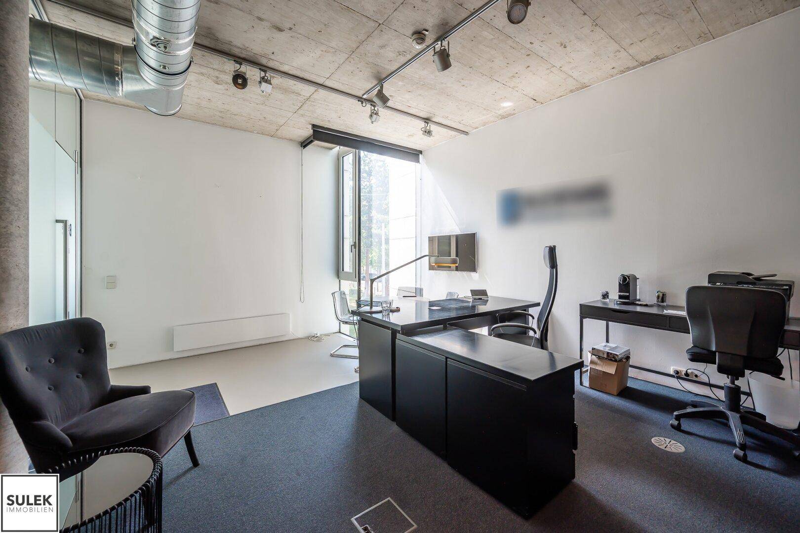 Weitläufiges Büro mit Schreibtisch, Bürostühlen, großem Fenster und grauem Teppichboden unter einer Sichtbetondecke.