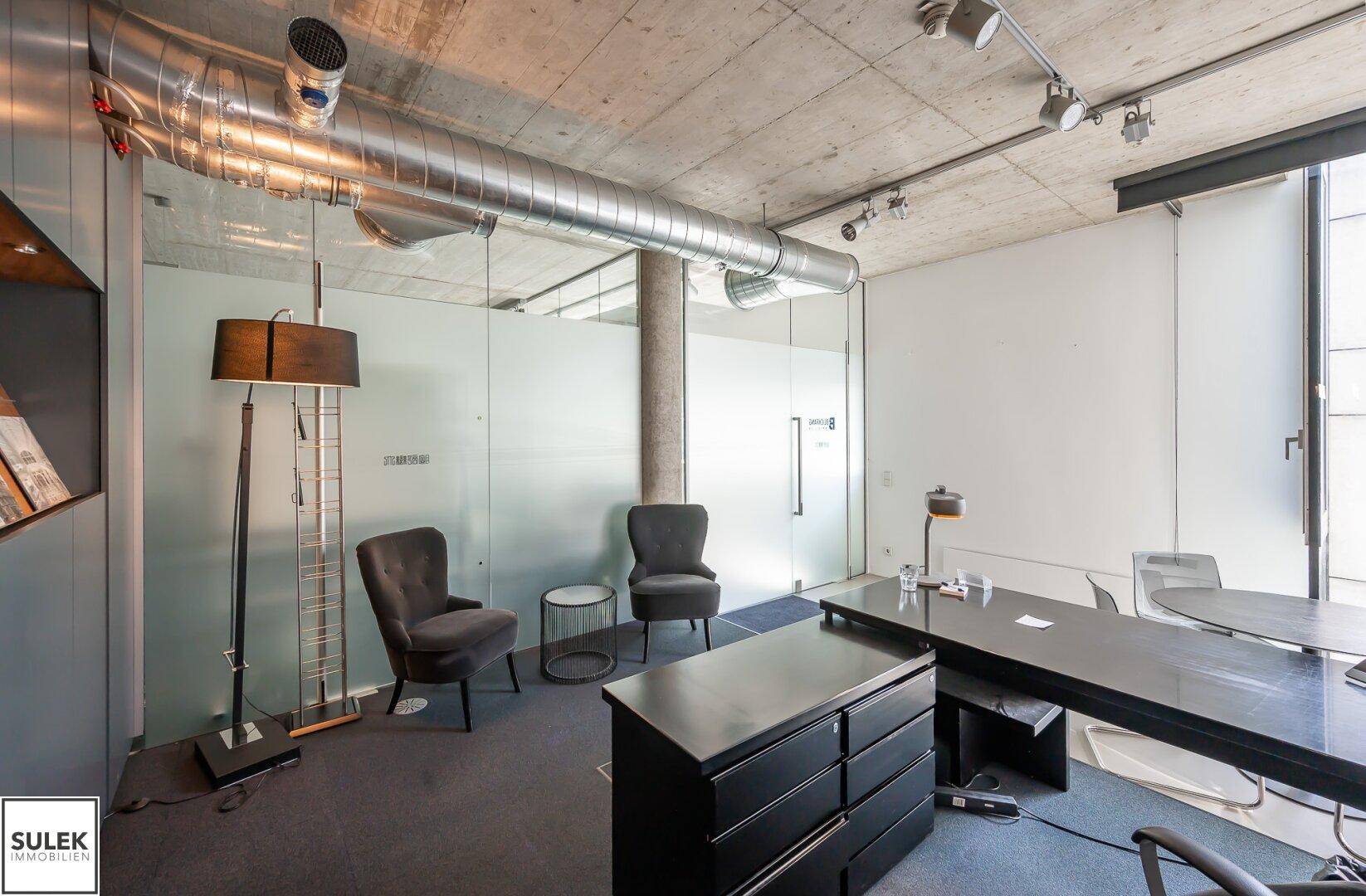 Geräumiges Büro mit Schreibtisch, bequemen Sesseln, Stehlampe und Glaswänden unter einer Sichtbetondecke.