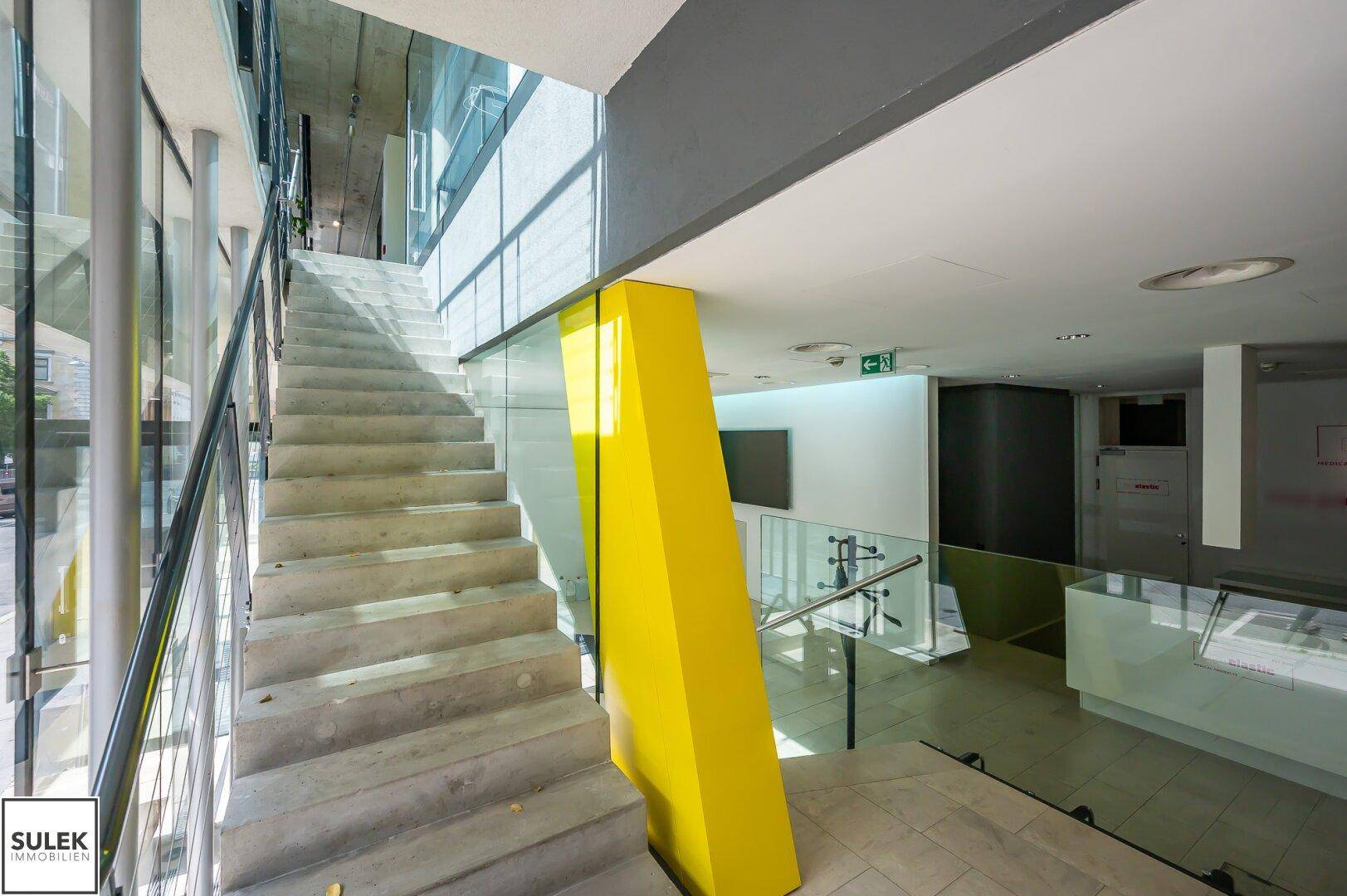 Moderne Treppe mit Betonstufen, Glasgeländer und einem auffälligen gelben Architekturelement, hell beleuchtet.