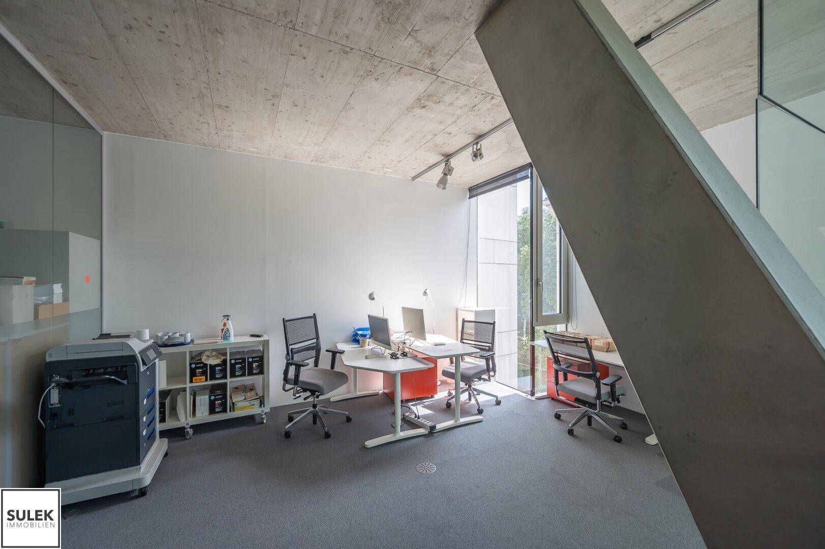 Geräumiges Büro mit mehreren Arbeitsplätzen, Sichtbetondecke und großen Fenstern für viel Licht.