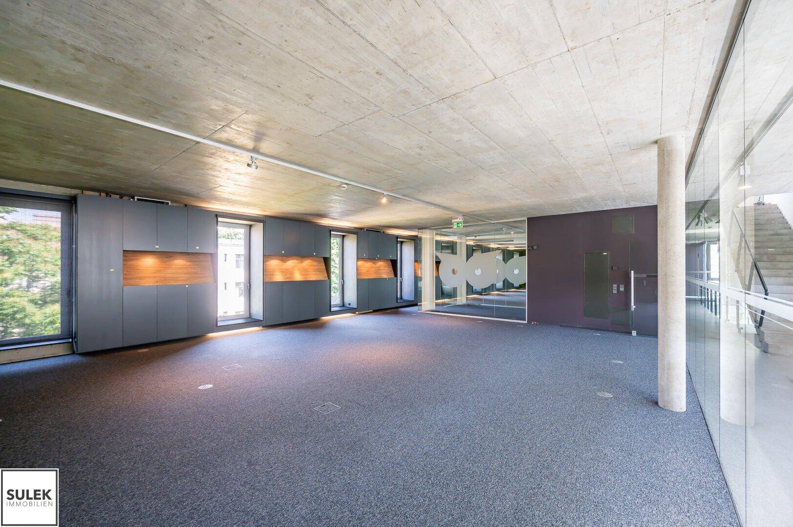 Geräumiger Bürobereich mit Teppichboden, unverputzter Betondecke und integrierten Schränken entlang der Fensterfront.