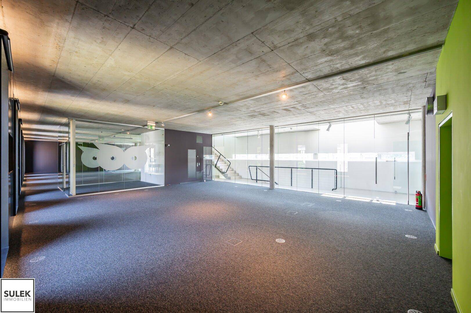 Heller Büroraum mit Teppichboden, Betondecke und Glaswänden, die den Blick auf den Treppenbereich freigeben.