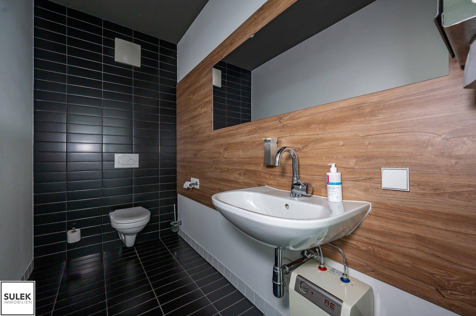Stilvolles Gäste-WC mit schwarzen Wand- und Bodenfliesen, Holzverkleidung und modernem Waschbecken.