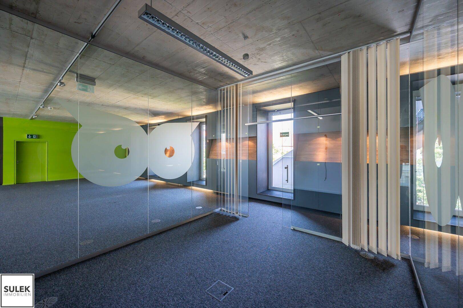 Moderner Büroraum mit Teppichboden, Betondecke und Glaswänden, die eine offene und helle Arbeitsumgebung schaffen.