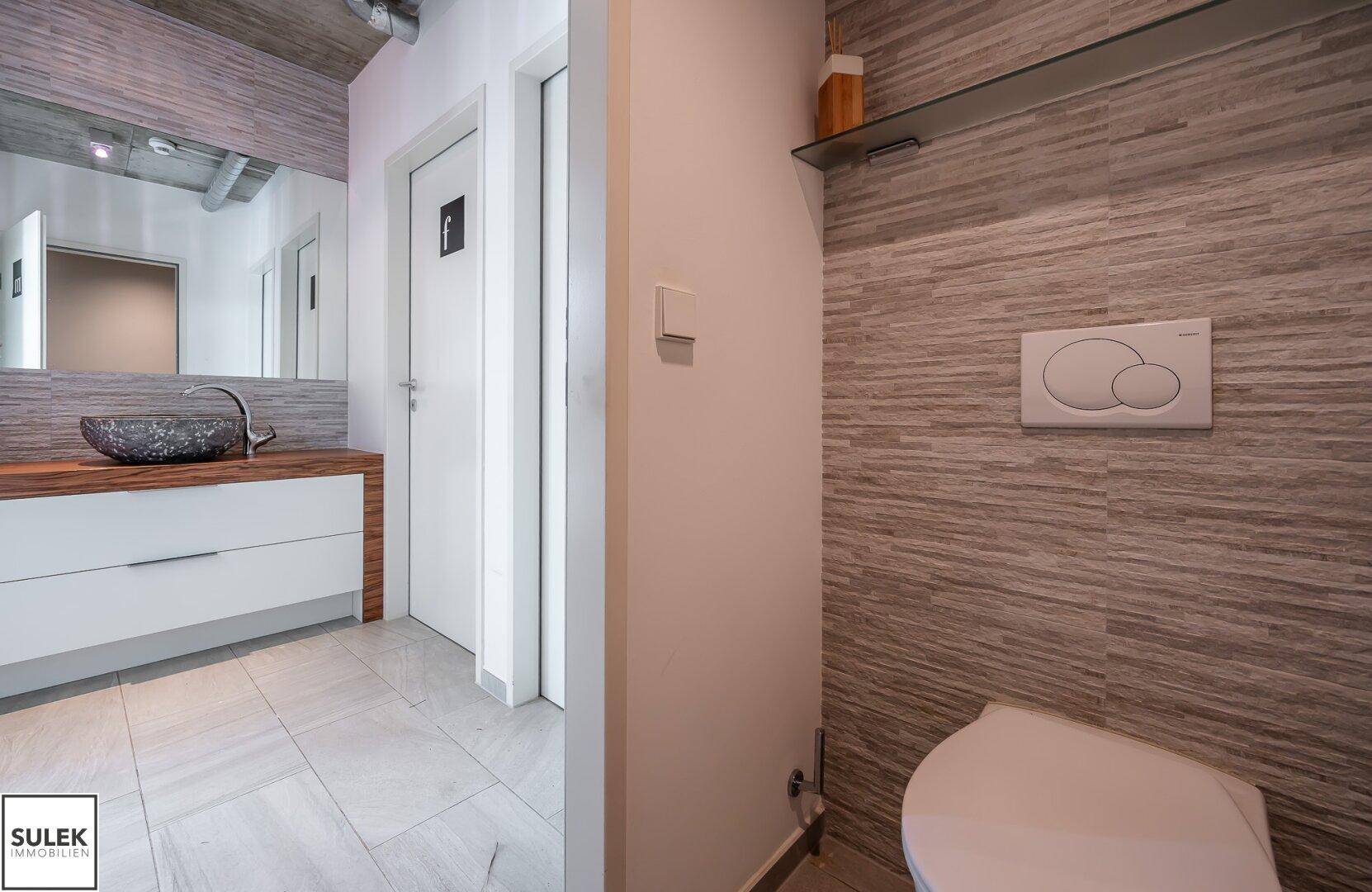 Modernes Badezimmer mit Steinwaschbecken, Holzunterschrank und gefliesten Wänden in Grautönen.