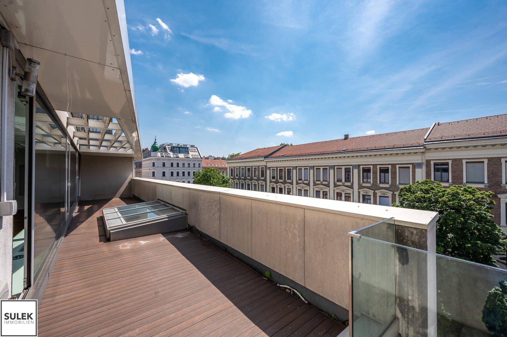 Geräumige Terrasse mit Holzboden, Glasgeländer und Blick auf die umliegenden Gebäude und Bäume.