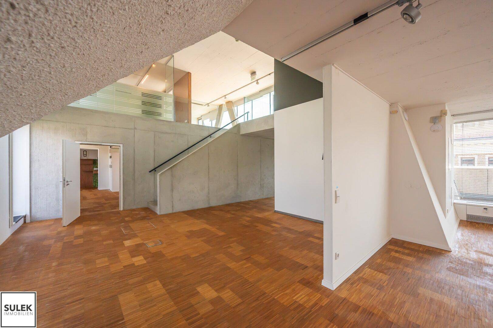 Geräumiger Innenraum mit Holzparkettboden, Betondecken und einer modernen Treppe mit Glasgeländer.