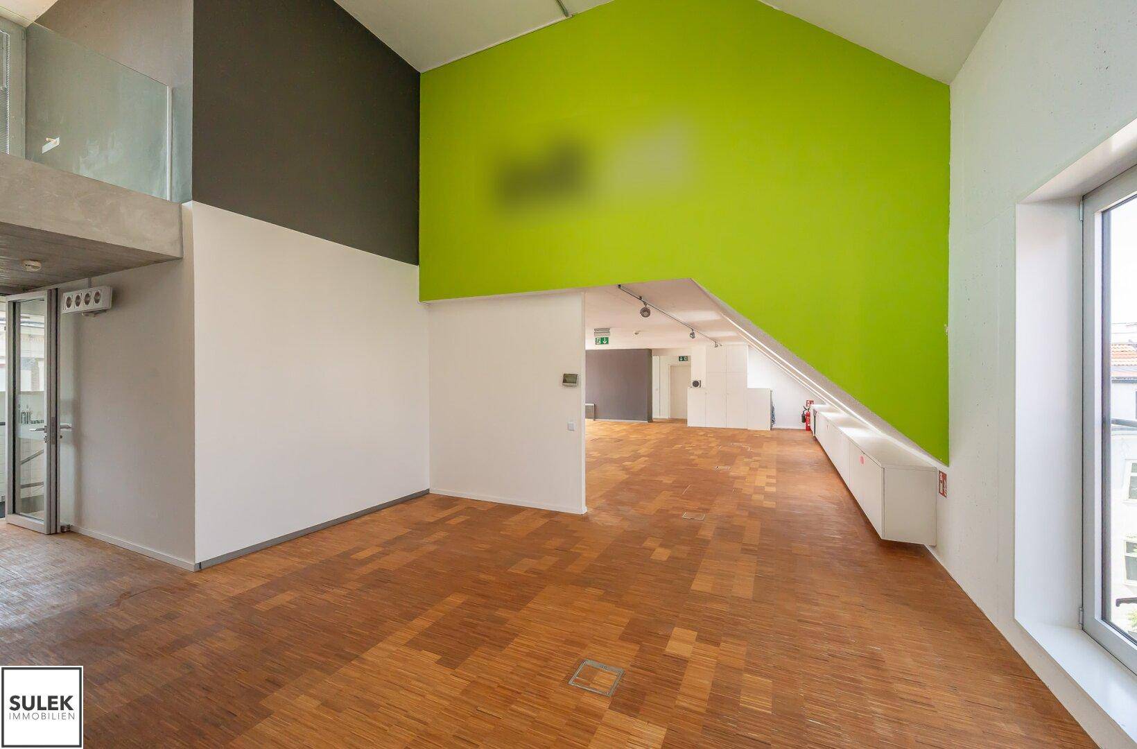 Offener Raum mit Holzparkettboden, einer leuchtend grünen Akzentwand und viel Tageslicht.