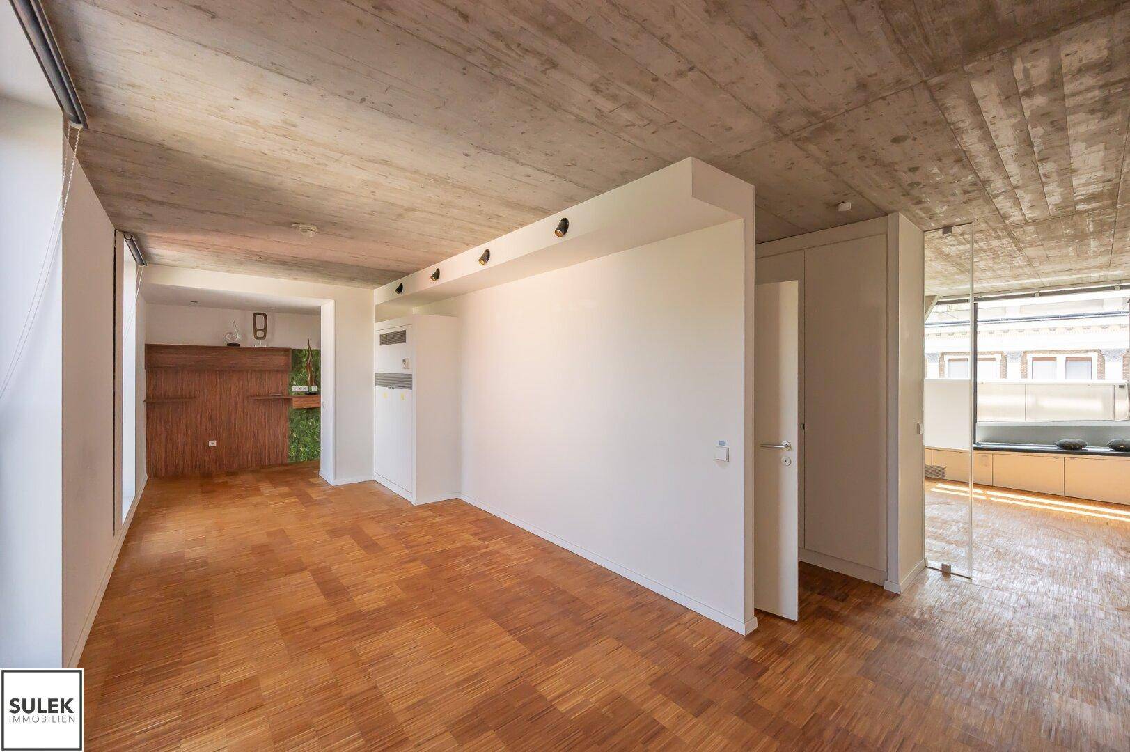 Offener Wohnbereich mit Holzparkettboden, freiliegender Betondecke und Blick auf die Küche.