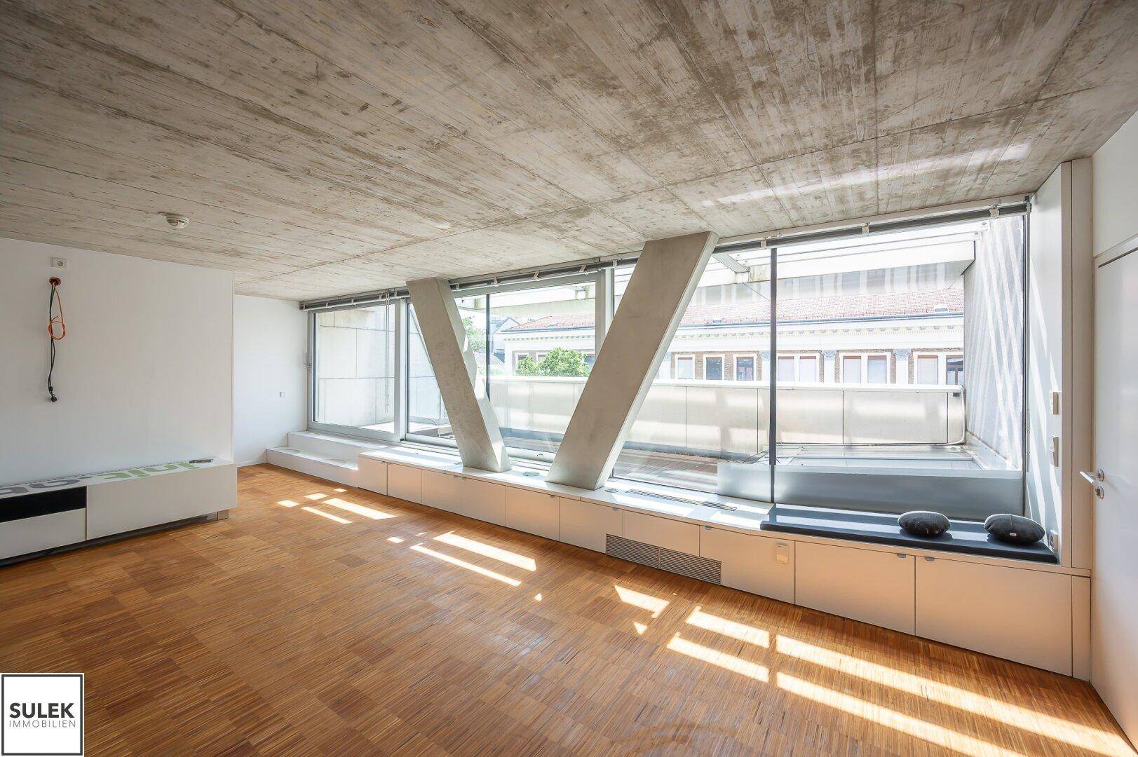 Heller Raum mit Holzparkettboden, Betondecke und einer Fensterfront mit integrierten Sitzgelegenheiten.