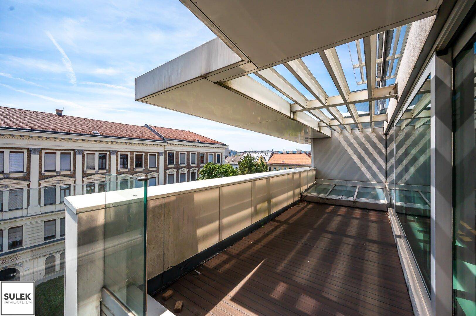 Geräumige Terrasse mit Holzboden, Glasgeländer und Blick auf die umliegenden Gebäude und Bäume.