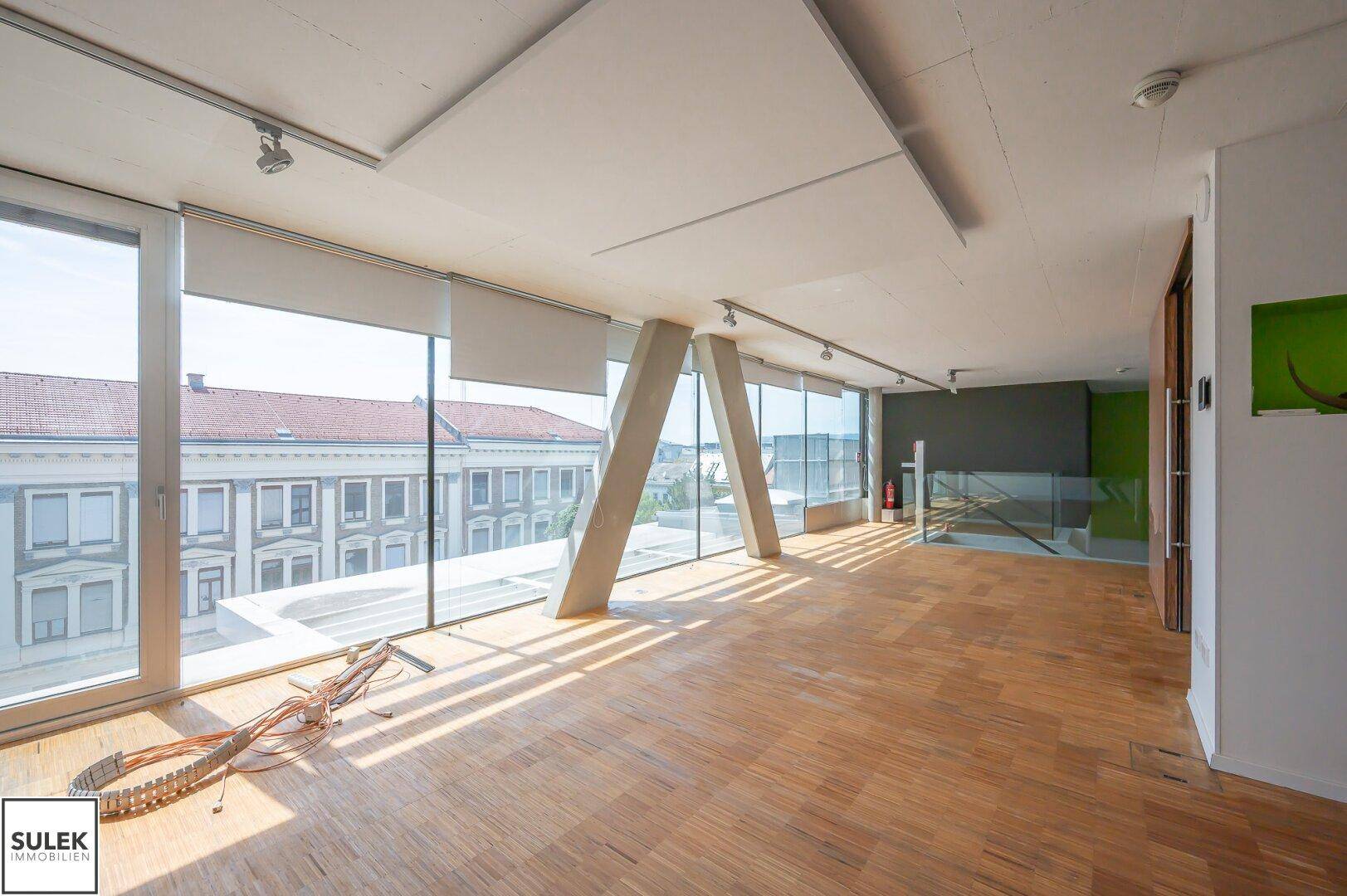 Großer, heller Raum mit Holzparkettboden, bodentiefen Fenstern und Blick auf die Stadt.