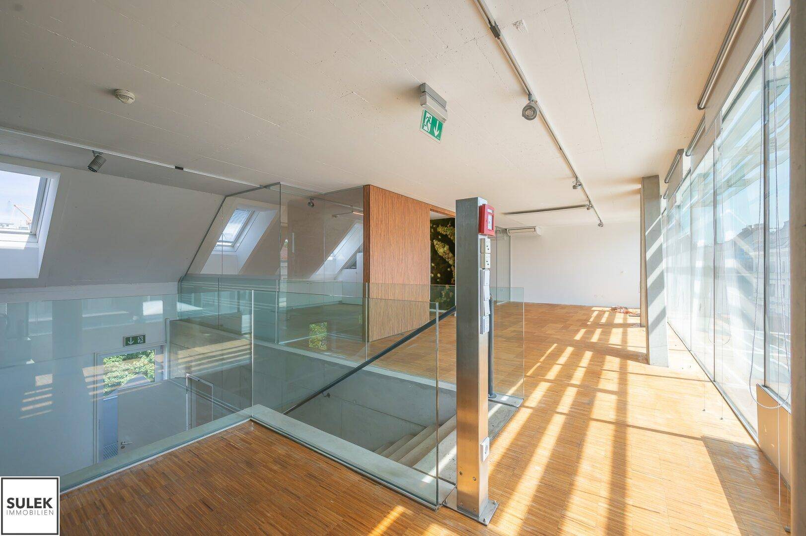 Heller Bürobereich mit Holzboden, Glasgeländer und Dachfenstern, die viel Tageslicht hereinlassen.