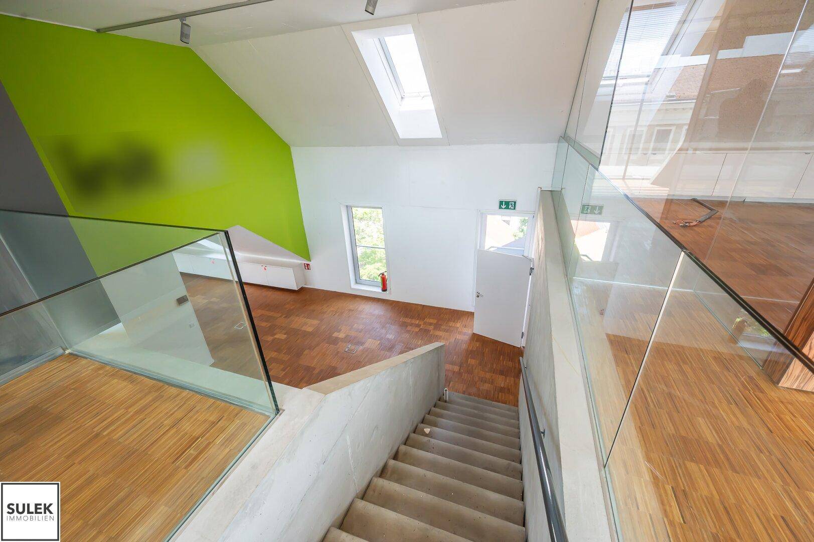Treppenhaus mit Glasgeländer und Holzboden, führt zu einem hellen Bereich mit grüner Wand.