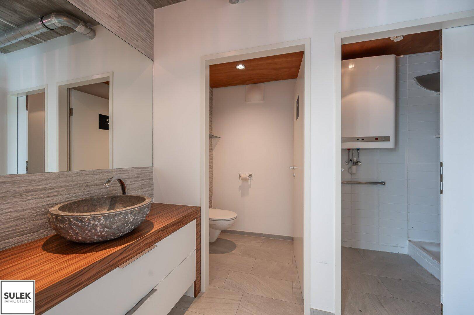 Modernes Badezimmer mit Steinswaschbecken, Holzunterschrank und separater Toilette, gut beleuchtet.