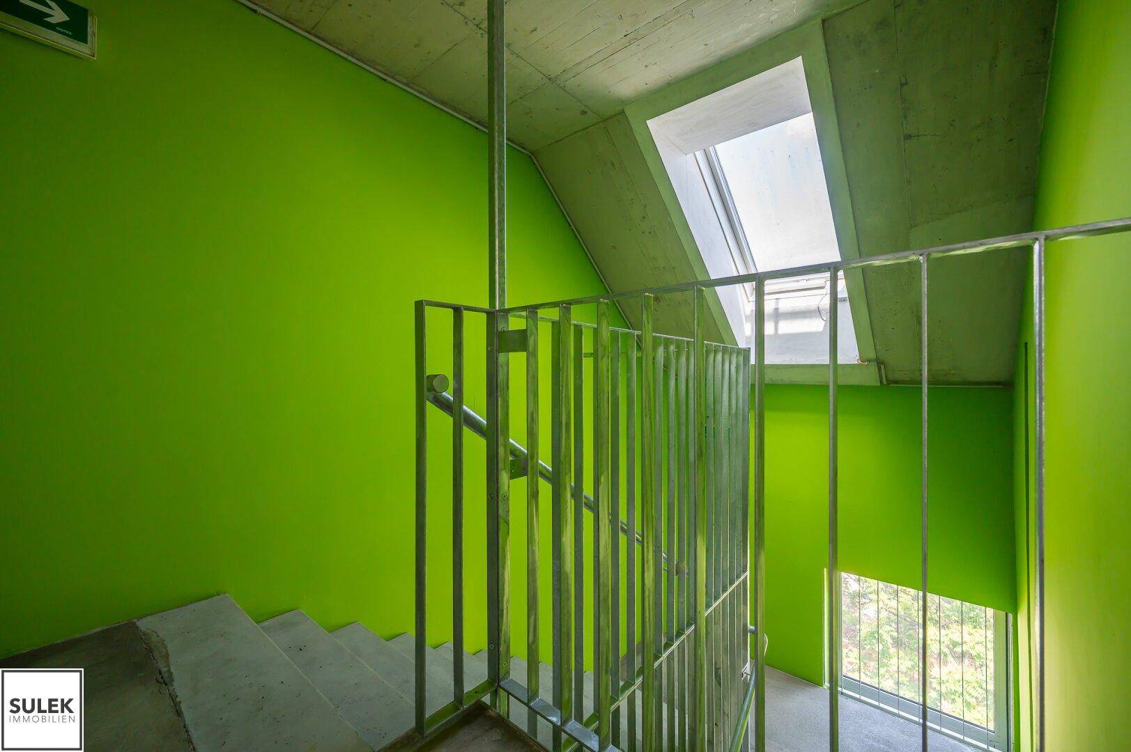 Treppenhaus mit leuchtend grünen Wänden und Metallgeländer, hell erleuchtet durch ein Dachfenster.