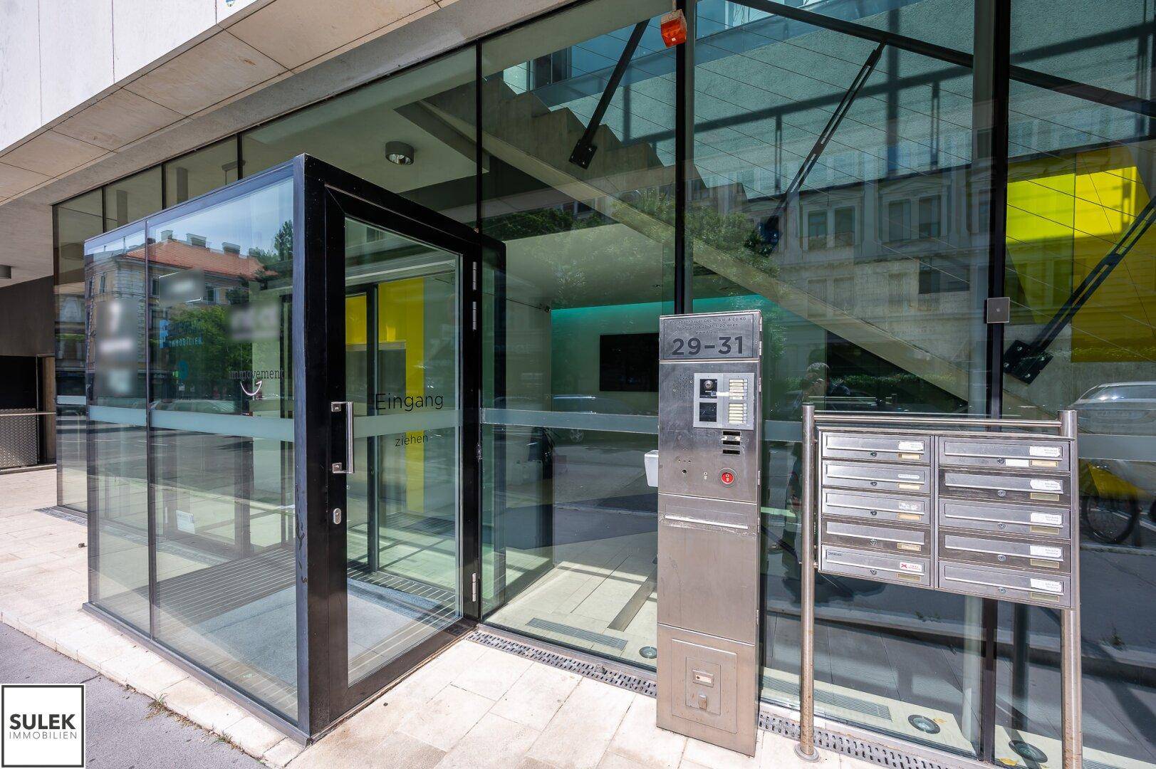 Eingangsbereich des Gebäudes mit gläserner Drehtür, Briefkästen und Gegensprechanlage unter hellem Tageslicht.