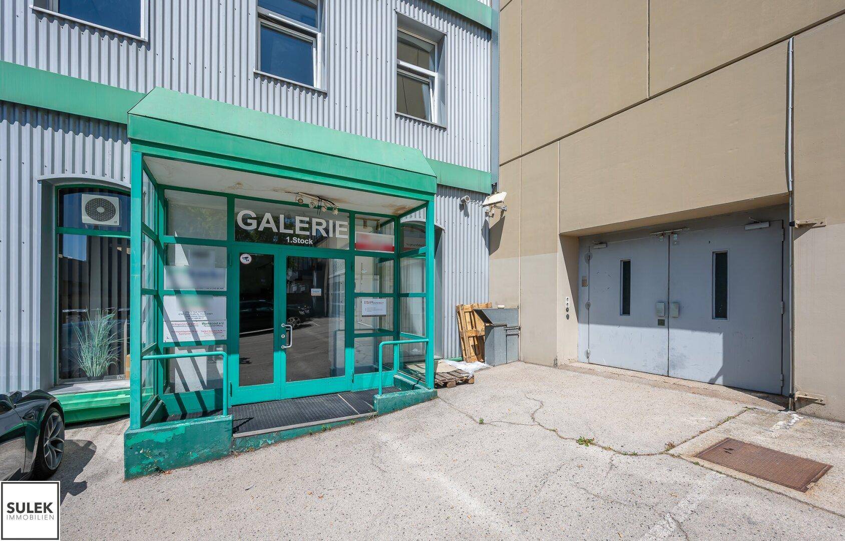 Eingangsbereich einer Galerie mit grüner Überdachung und Glastüren, daneben ein weiterer Zugang.