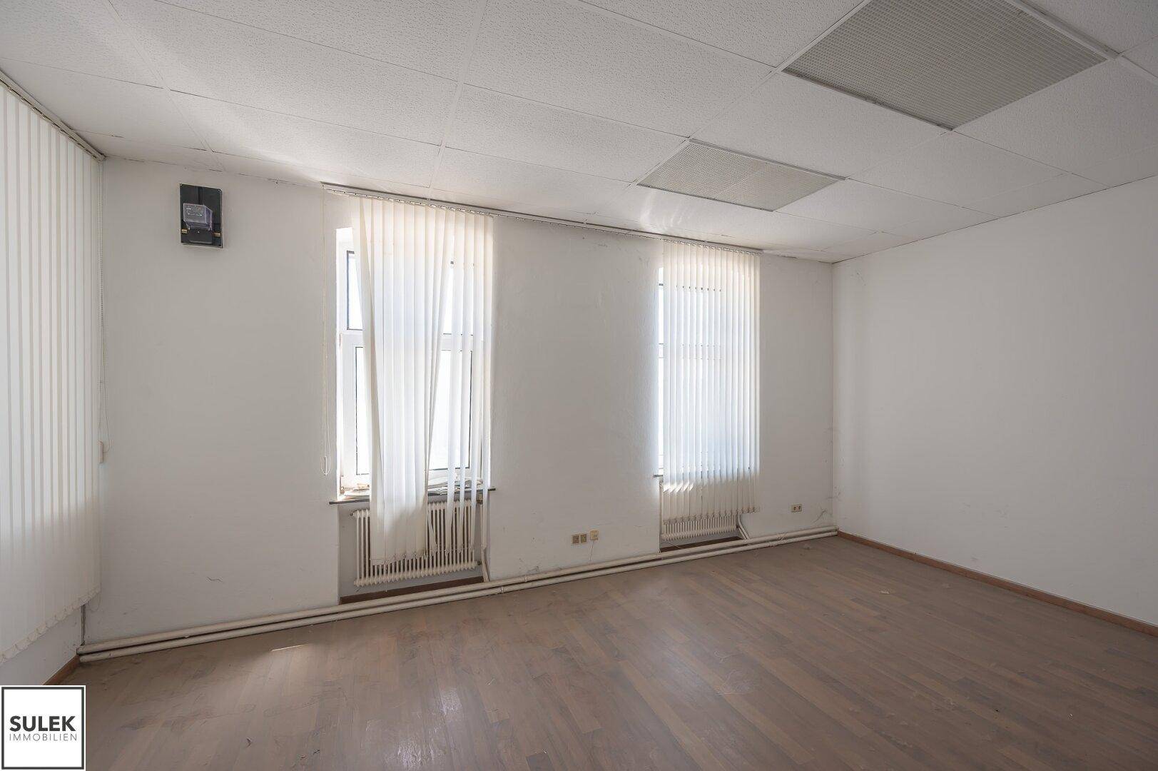 Leerer Büroraum mit zwei Fenstern, weißen Wänden und einem Holzboden, ausgestattet mit Heizkörpern.
