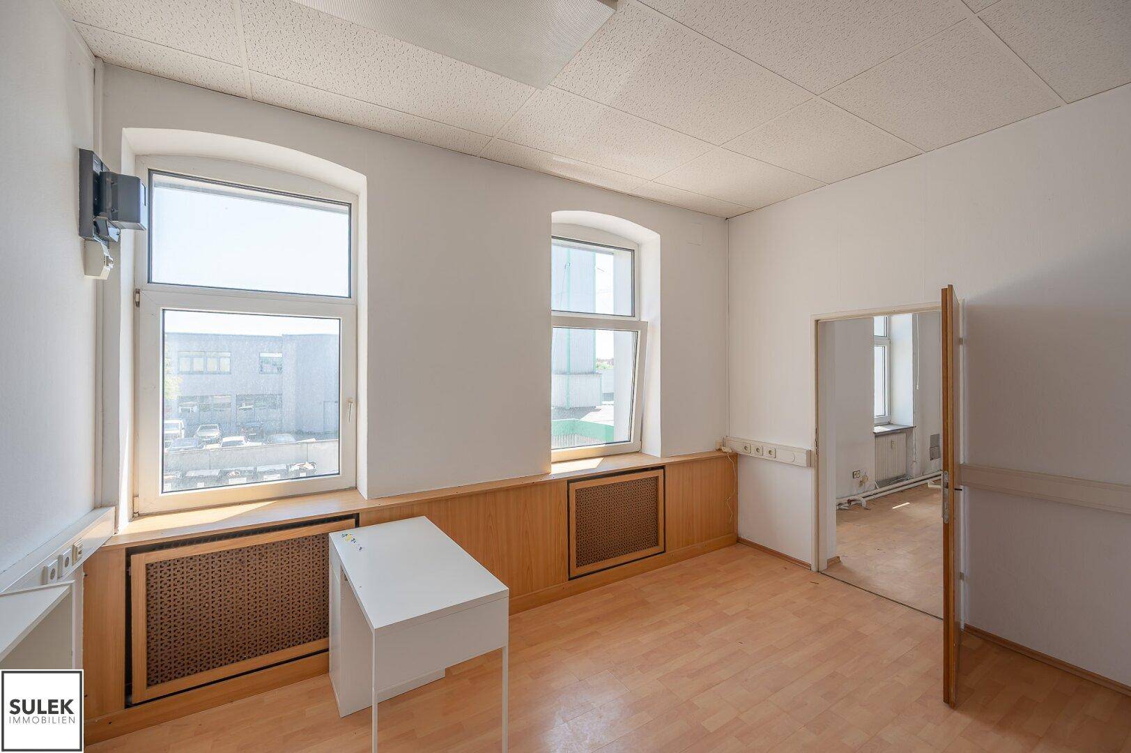 Leerer Büroraum mit Holzboden und Holzverkleidung unter den Fenstern, ein kleiner Tisch steht im Raum.