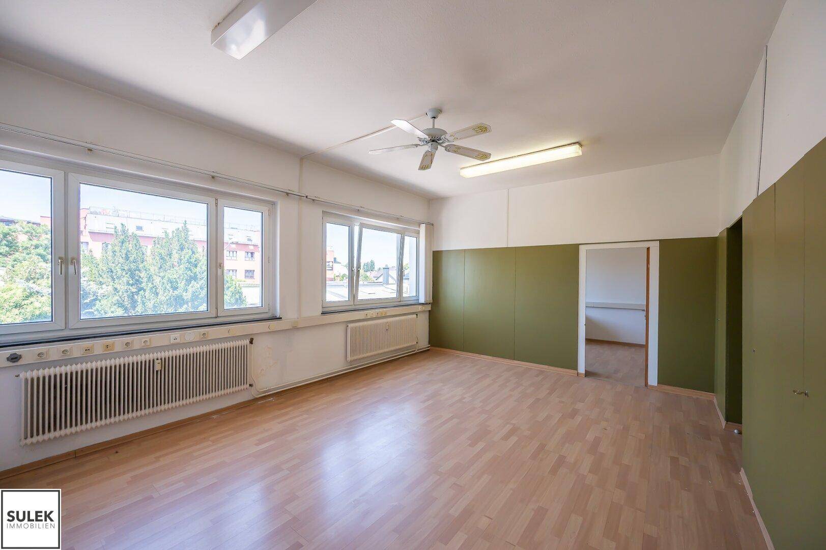 Geräumiger Büroraum mit vielen Fenstern, Holzboden und einem Deckenventilator, die Wände sind zweifarbig gestaltet.