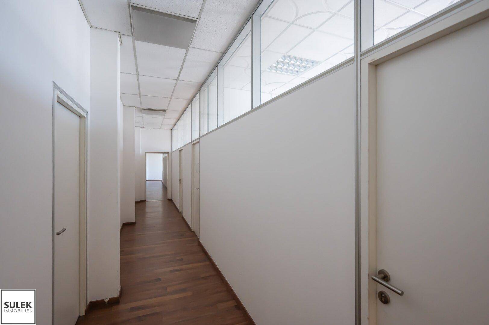 Langer Flur in einem Bürogebäude mit Holzboden und Glasabtrennungen zu den angrenzenden Räumen.