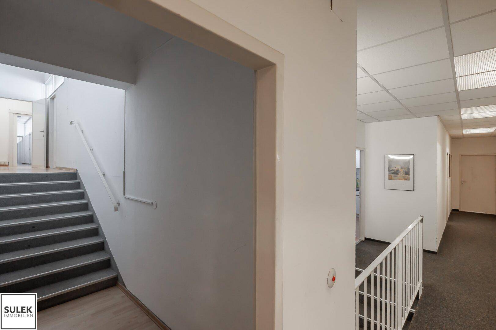 Treppenhaus mit grauem Teppich und weißem Geländer, das zu einer weiteren Etage führt, daneben ein Flurbereich.