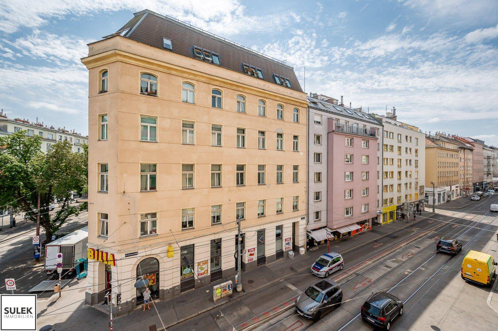 Panoramablick auf eine belebte Stadtstraße mit einem markanten Eckgebäude und städtischer Architektur.