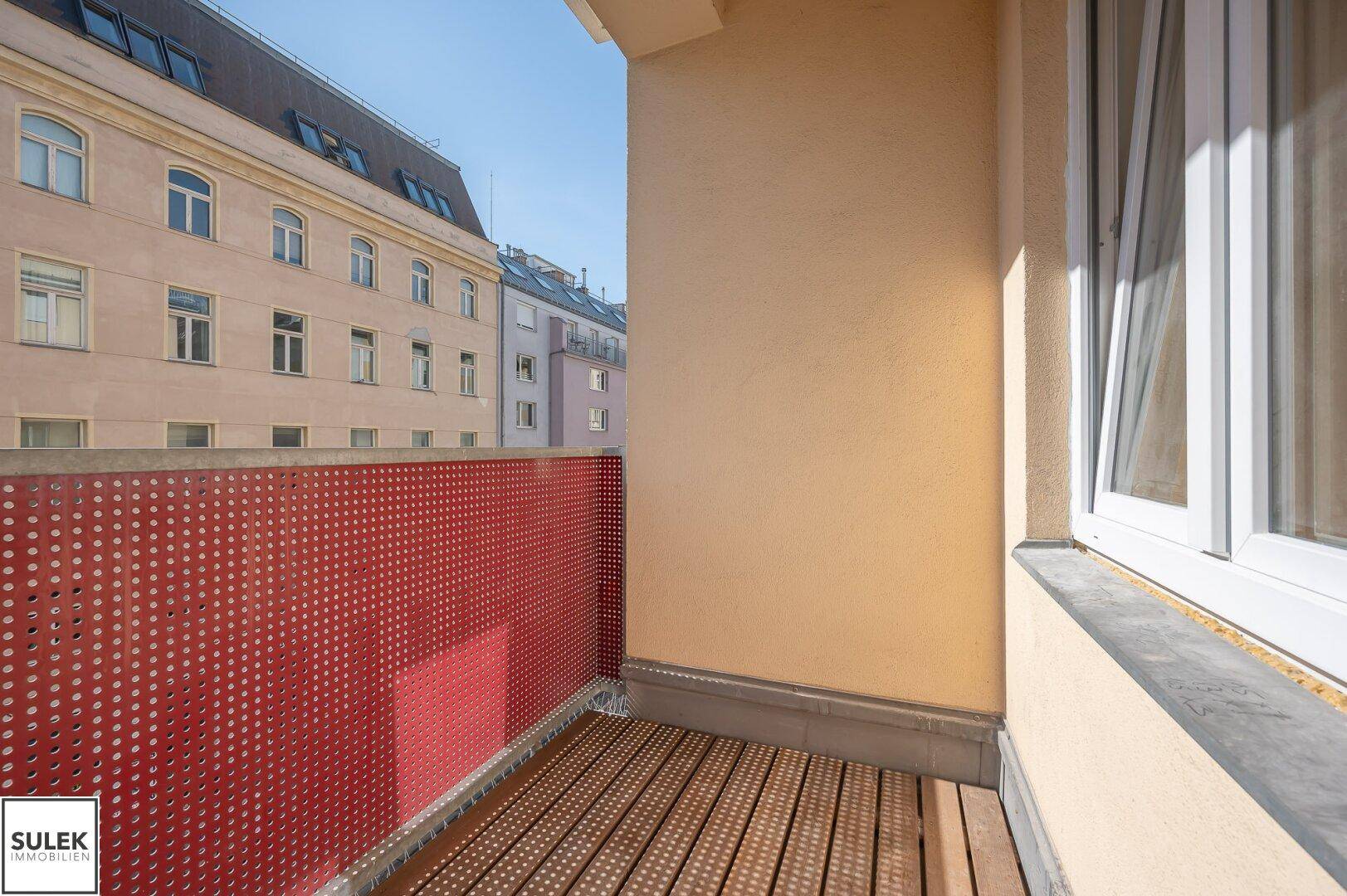 Kleiner Balkon mit Holzboden und rotem Geländer, bietet einen Blick auf die umliegenden Gebäude.