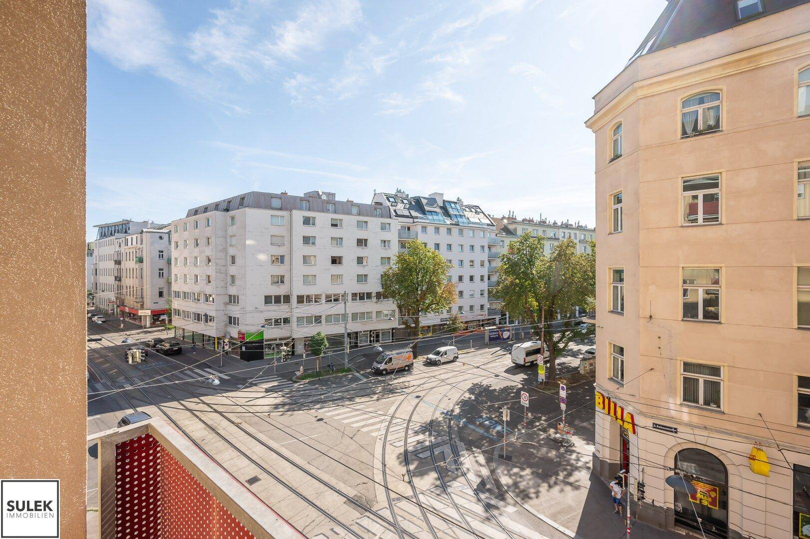 Weitblick über eine belebte Stadtkreuzung mit Straßenbahnschienen und umliegenden Wohngebäuden.