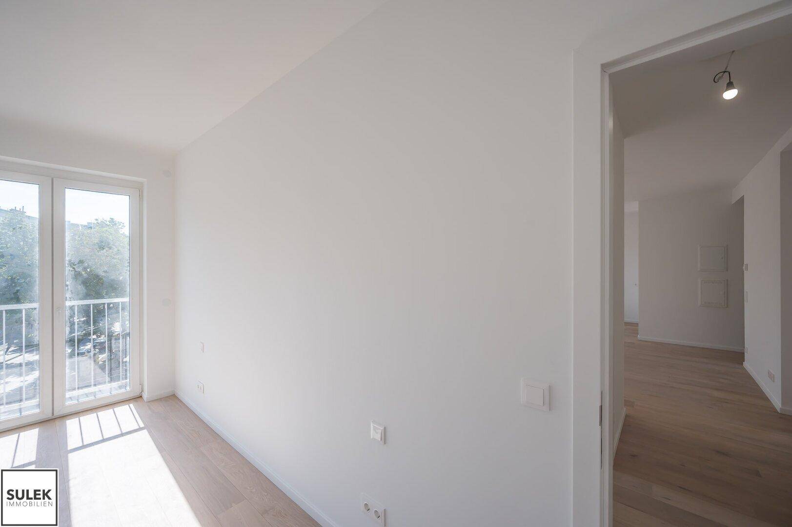 Lichtdurchflutetes Zimmer mit Holzboden, Balkonzugang und offenem Übergang zu einem weiteren Raum.