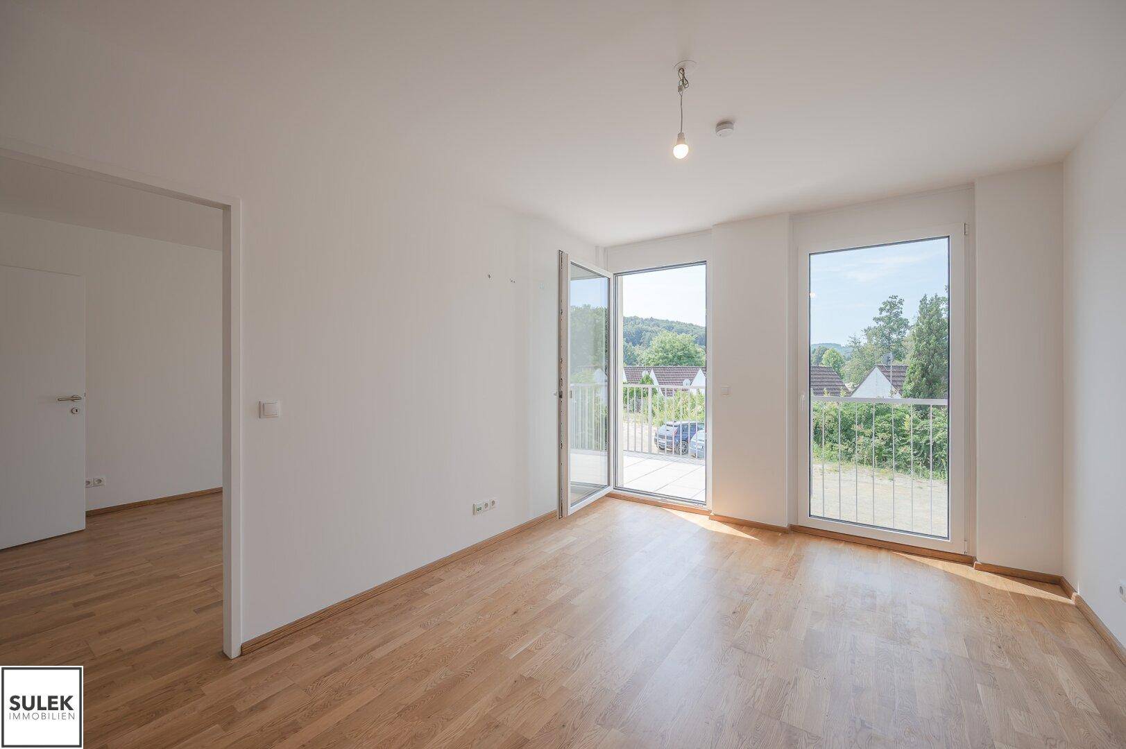 Lichtdurchfluteter Raum mit Holzboden und zwei Fenstern, die Zugang zum Balkon bieten.