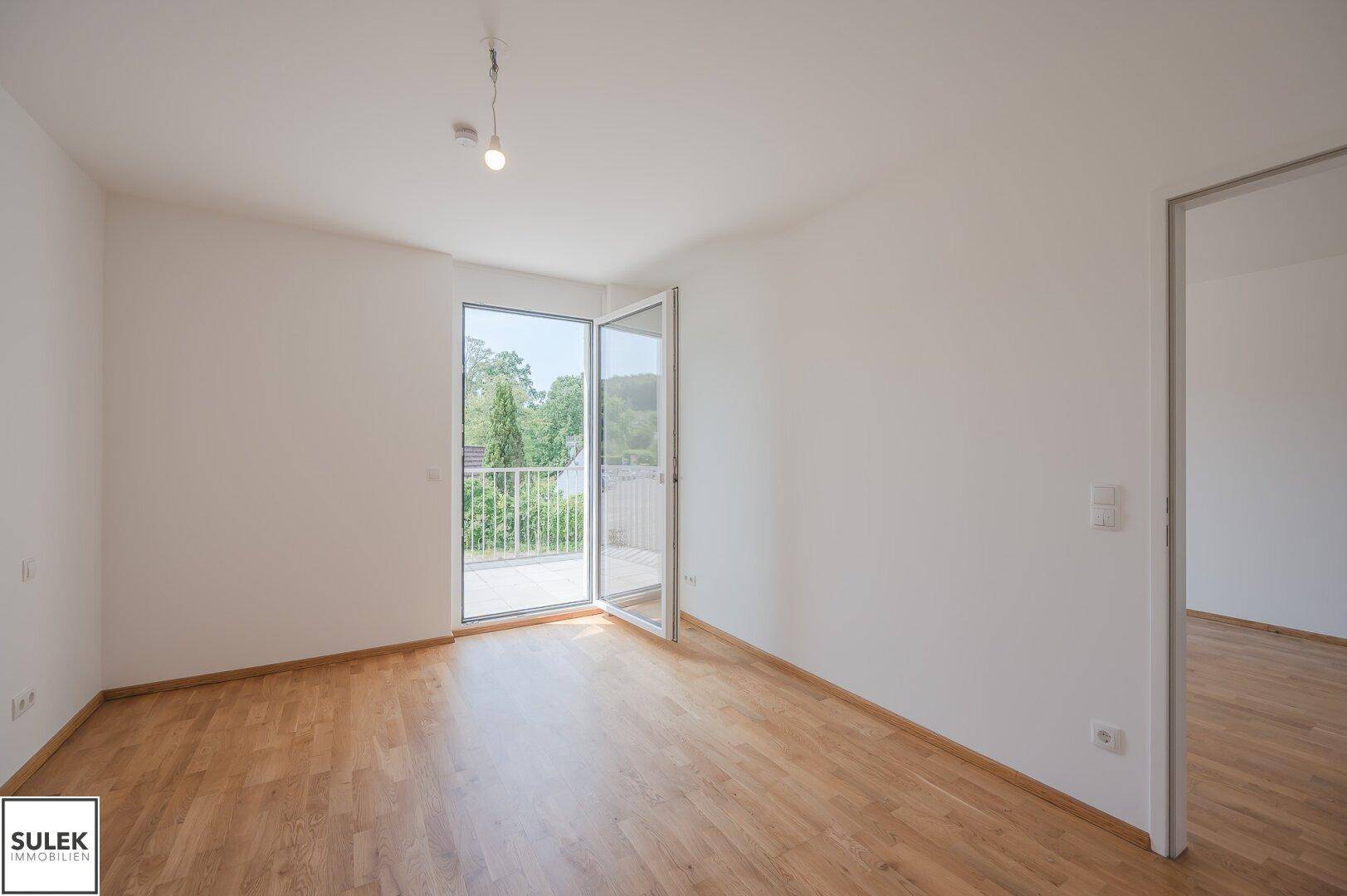 Helles Wohnzimmer mit Holzboden und großem Fenster mit Zugang zum Balkon.