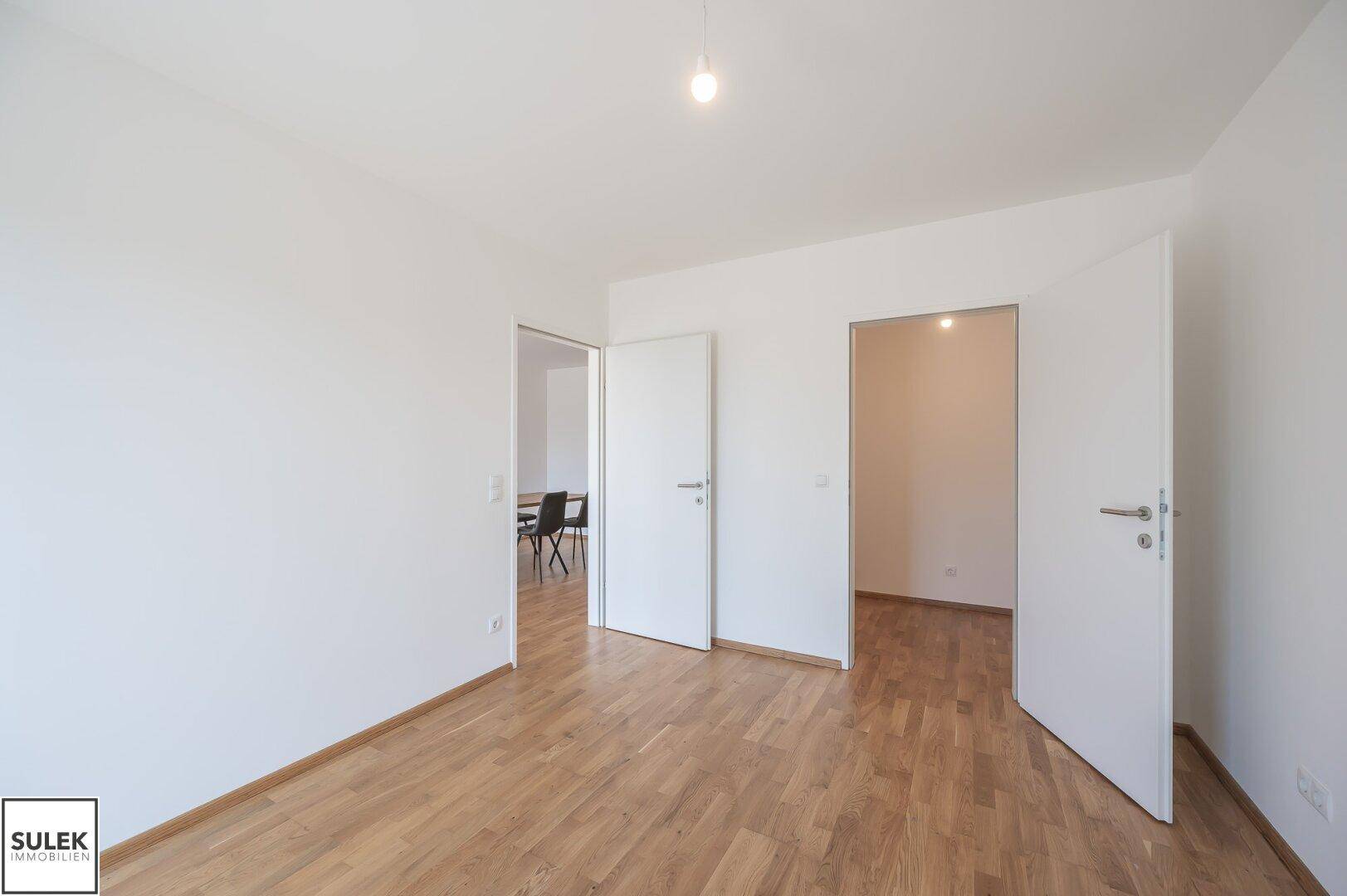 Heller Raum mit Holzboden und weißen Wänden, der zu weiteren Zimmern führt.