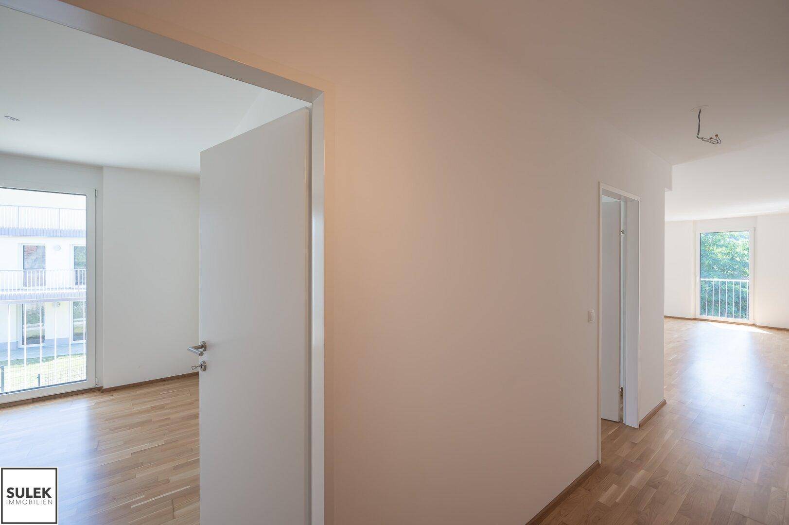 Heller Flur mit weißer Tür und Blick in einen angrenzenden Raum mit Holzfußboden und Fenster.